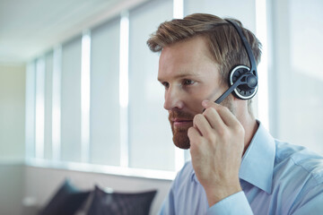 Male office worker wearing dress shirt adjusting headset mic at office desk with blinds, copy space