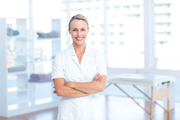 Female therapist in white uniform smiling in therapy room with table and shelves with towels