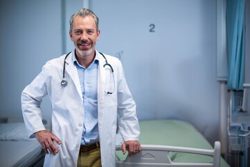 Male doctor wearing lab coat leaning on bed rail in hospital ward with stethoscope, privacy curtain