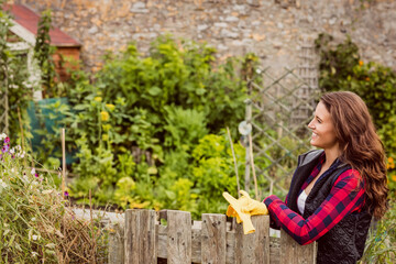 Woman leaning on picket fence in garden with plants holding yellow gardening gloves, copy space