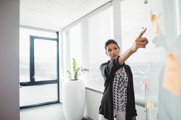 Asian woman in business casual pointing at sticky notes on glass partition near planter in office