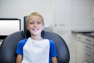 Child patient sitting on reclining dental chair in treatment room wearing protective bib by monitor