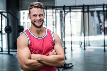 Muscular male standing near squat rack, large windows in gym, wearing red tank top, copy space