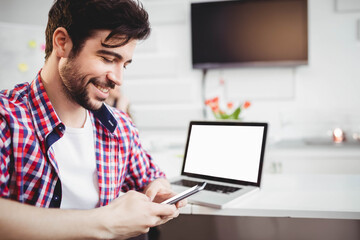 Man smiling while checking smartphone at home workspace with laptop and tulip vase, copy space