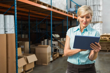 Middle-aged woman holding tablet while checking stock on shelving in warehouse, copy space