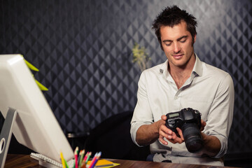 Man examining DSLR camera while sitting at wooden desk with computer monitor and colored pencils