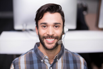 Support agent wearing plaid shirt and headset is smiling at call center desk with monitors behind
