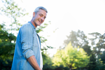 Man standing in lush sunlit park looking over shoulder and smiling under green canopy, copy space