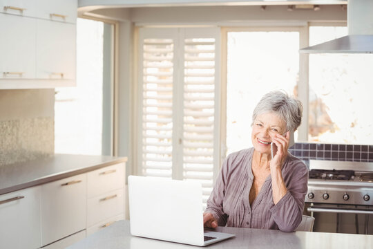 Senior woman using smartphone and typing on laptop at kitchen island near windows