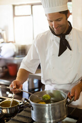 Male chef wearing uniform stirring green vegetables in steel pot on gas stove in restaurant kitchen
