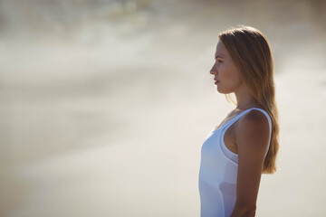 Woman wearing white tank top standing at shoreline, facing right and watching waves, copy space