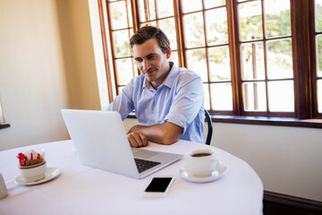 Man wearing button-down shirt typing on laptop at round white table in bright room with smartphone