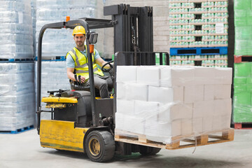 Male forklift operator wearing helmet operating forklift in warehouse carrying boxes on pallet