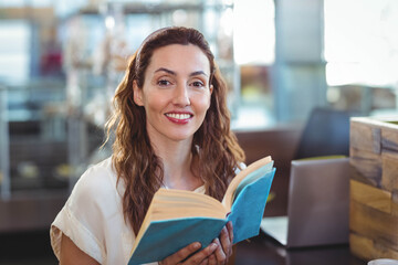 Woman reading blue-bound book while sitting at cafe table by partition with silver laptop nearby