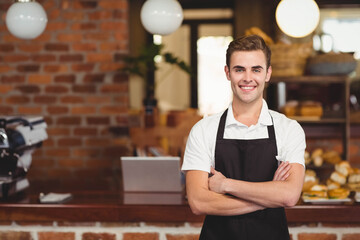Male barista wearing apron behind cafe counter crossing arms near espresso machine, copy space