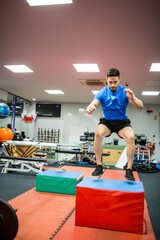 Male athlete jumping over red foam plyometric box at gym with benches, weights, racks, and mats
