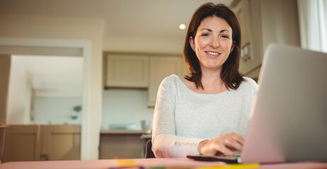 Woman typing on laptop at bright kitchen workspace with light wood cabinets behind, copy space