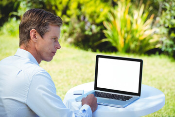 Laptop sitting on round plastic table on lawn near garden shrubs, with notepad, pen and smartphone