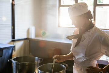 Chef wearing white coat and hat stirring pot on stovetop as sunlight streaming through windows