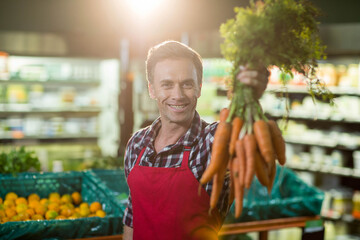 Male grocery worker wearing red apron holding bunch of carrots toward camera in produce aisle