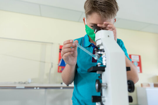 Boy wearing green mask peering into white microscope holding pipette and slide at classroom bench