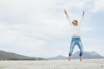 Obraz premium Woman jumping on sandy plain under sky wearing button-down shirt and rolled-up jeans, copy space