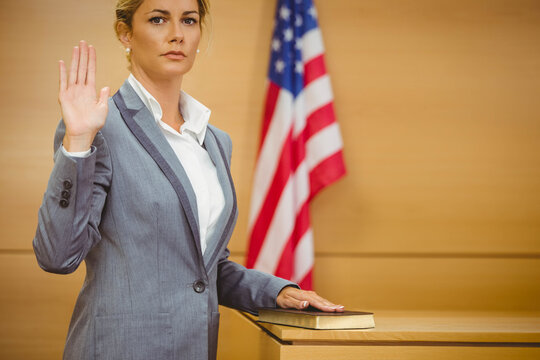 Woman standing in witness stand raising right hand on Bible beside US flag in grey suit