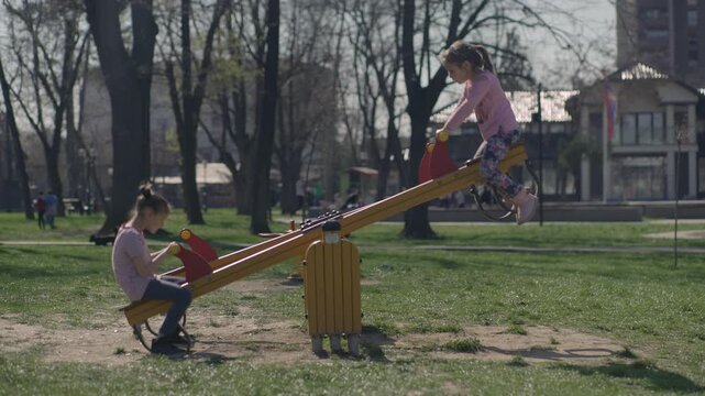 Little girls swinging on the seesaw in the park, trees in the blurred background, spring season.