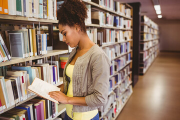 Woman reading open book in library aisle, with tall metal bookshelves under fluorescent lighting