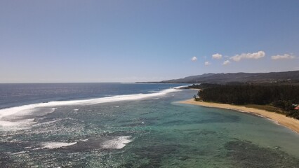 Aerial drone view of the tropical coastline in Mauritius with waves, beach, forest, and distant mountains on a sunny day.