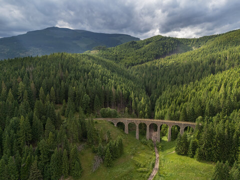 Aerial view of a stone bridge cutting through the dense, emerald forest under a brooding sky, casting shadows and highlights across the landscape, Telg&Atilde;&iexcl;rt, Brezno District, Slovakia.