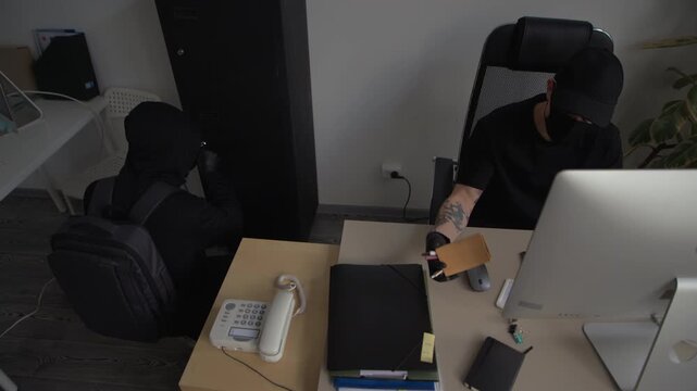 High angle shot of robber trying to break into safe when male accomplice hacking into corporate computer then giving key to partner during industrial espionage in office
