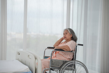 Depressed senior Asian woman patient in hospital sitting in wheelchair by the window, upset...