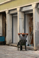 worker resting on plastic chair while colleague paints building facade