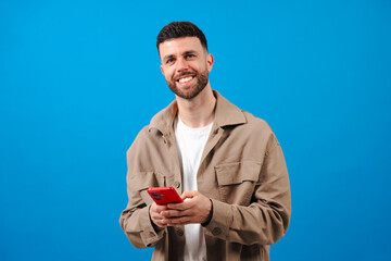 Portrait of a cheerful young bearded man in a beige jacket and whit shirt, holding a red smartphone and smiling confidently against a solid blue studio backdrop, symbolizing modern digital