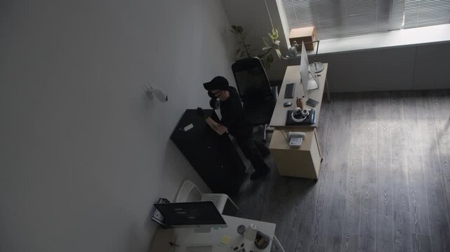 High angle shot of masked male criminal blocking surveillance camera on wall with adhesive tape blinding security system then hacking into computer while spying in corporate office