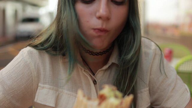 Turquoise-haired teenager girl biting street burrito, sipping drink while seated at urban cafe during bright sunlight with casual, relaxed demeanor