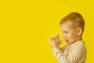 A cute boy drinks fresh water from a clear glass against a yellow wall. Drinking water for children.