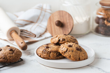 Stack of fresh chocolate American style chip cookies with nuts and chocolate drops on a tray with transparent lid on white marble background. Cookies inside a glass jar with kitchen utensils
