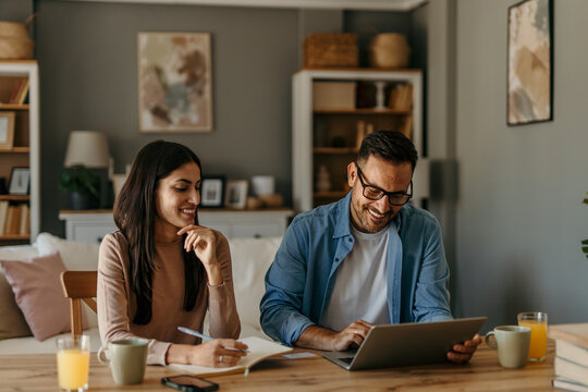 Couple working from home using laptop and taking notes smiling at each other
