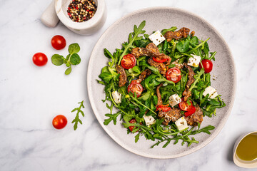 Warm beef meat salad with arugula, lettuce, feta and tomatoes on a plate on a light stone background, top view. A delicious gourmet dinner in the rustic style. High quality photo