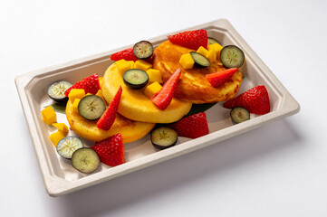 Fluffy pancakes topped with fresh strawberries, mango, kiwi, and blueberries, served in a takeout container on a white table. Natural light highlights this healthy, colorful breakfast or light snack.P