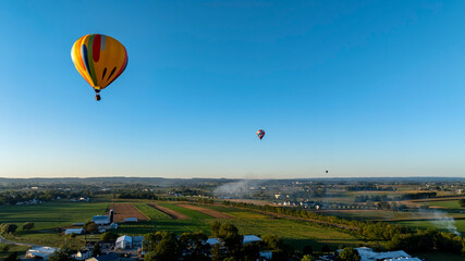 Obraz premium Hot air balloons gracefully rise against a clear blue sky, gliding over expansive green fields and farms during the early morning hours. The tranquil landscape showcases the beauty of nature.