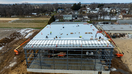 Workers are busy installing a roof on a large commercial building under construction in a rural area. The surrounding landscape shows houses and fields, with snow visible on the ground. © Greg Kelton