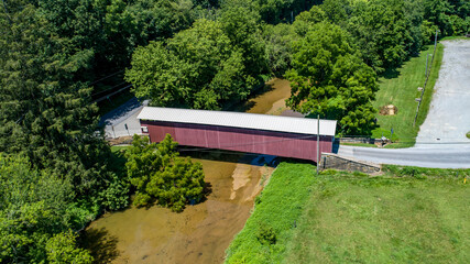 A serene covered bridge spans a calm stream, surrounded by dense greenery and open grassland. This picturesque scene captures the beauty of nature on a sunny day, inviting exploration.