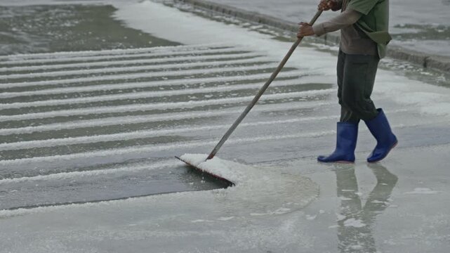 Man in rubber boots walks and picks up salt pile along evaporation pond closeup. Worker gathers salty mineral deposit to dry with traditional tool at seaside quarry. Craft industry