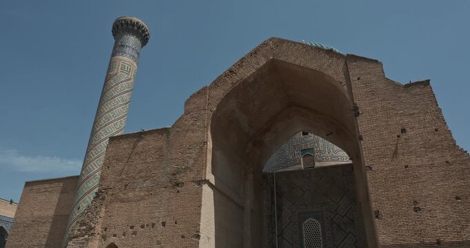 Ancient ruins of brick entrance wall in front of turquoise dome and tall minaret of Gur e Amir Mausoleum in Samarkand, Uzbekistan under blue sky, showcasing Islamic architecture, restoration contrast