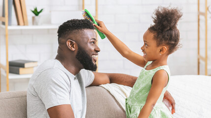 A young black girl with curly hair is brushing her fathers hair with a green brush while he sits on...