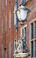 Ornate street lamp with sculpted iron flourishes stands before Gdansk’s historic brick facade —...