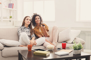 Smiling female friends relaxing and watching TV at home, changing channels, having rest after hard...
